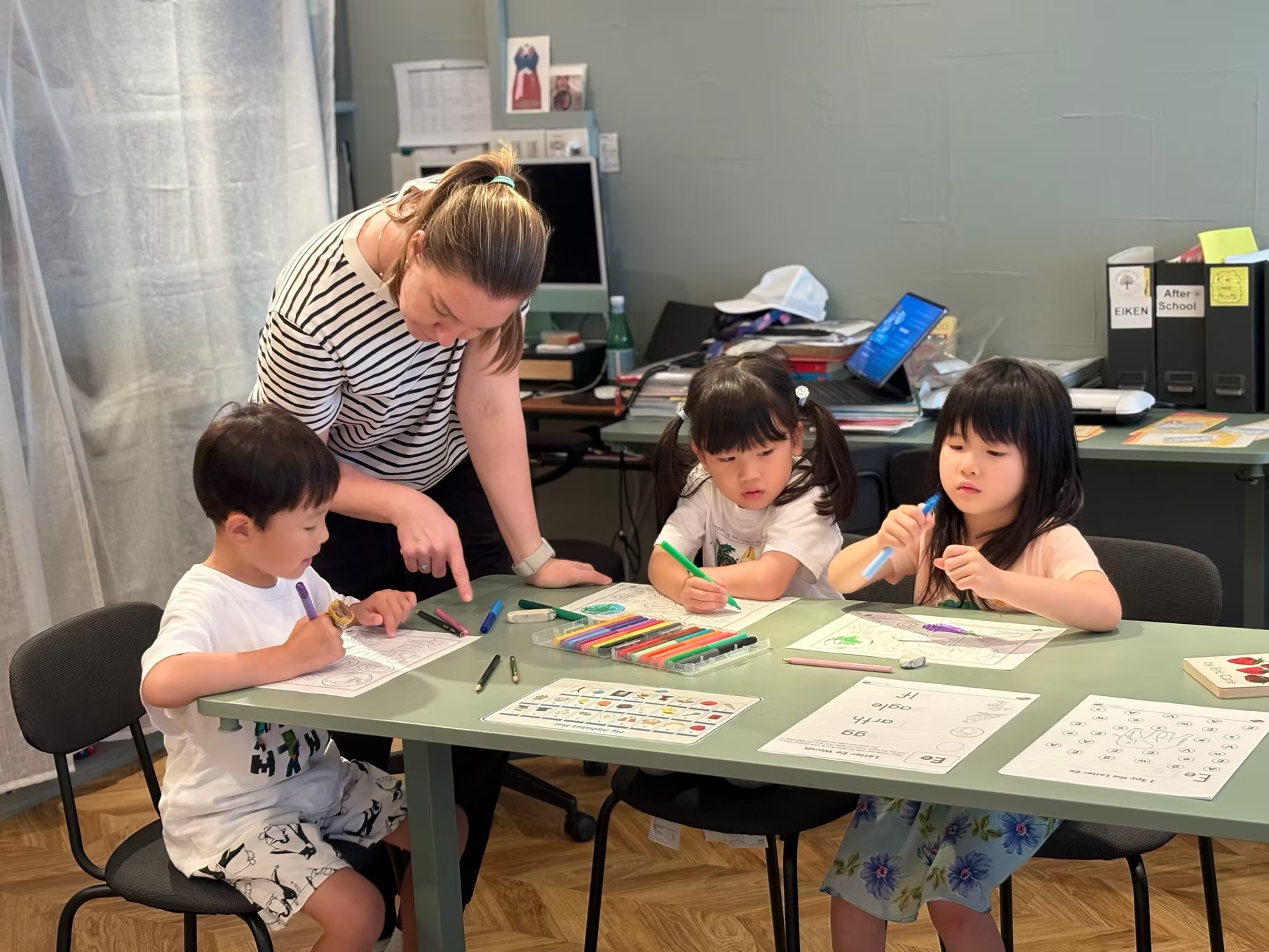 A teacher gently guides three kindergarten-age children as they draw and explore letters together at a classroom table.
